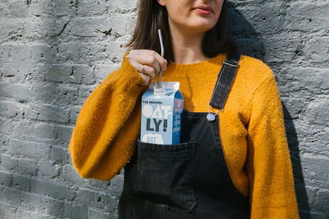 Kvinna iklädd orange stikad tröja stårmot stenvägg, i bröstfickan på hennes hängelbyxor har hon en blå tetra med Oatly med ett sugrör i. Bilden utstrålar brand love.